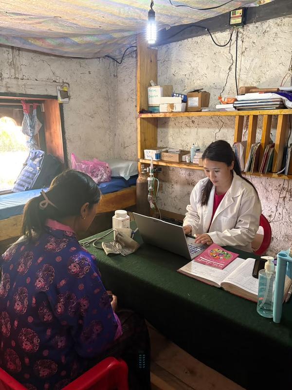 Dema, in a white coat, works at a laptop in a small health room while a woman sits across the desk from her.