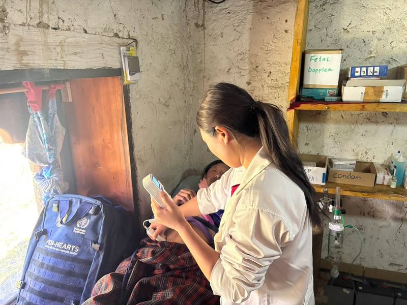 Dema examines a patient lying on a bed, holding a handheld monitor.