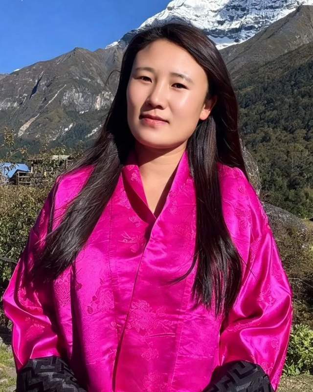 Dema in a pink silk jacket, standing outdoors with snow-capped mountains behind her.