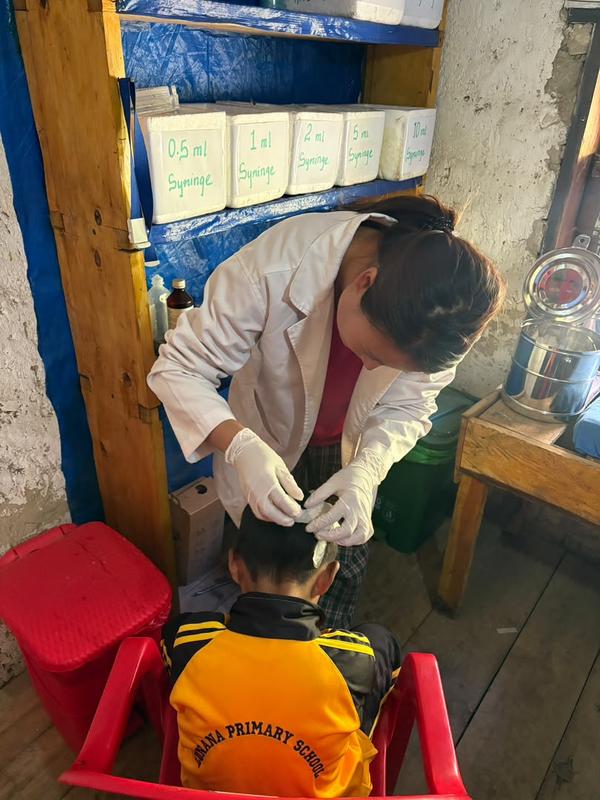 Dema bends over a schoolboy to treat his head, beside shelves of labelled syringe boxes.