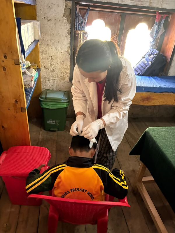 Dema, wearing gloves, tends to the head of a young schoolboy seated on a red chair.